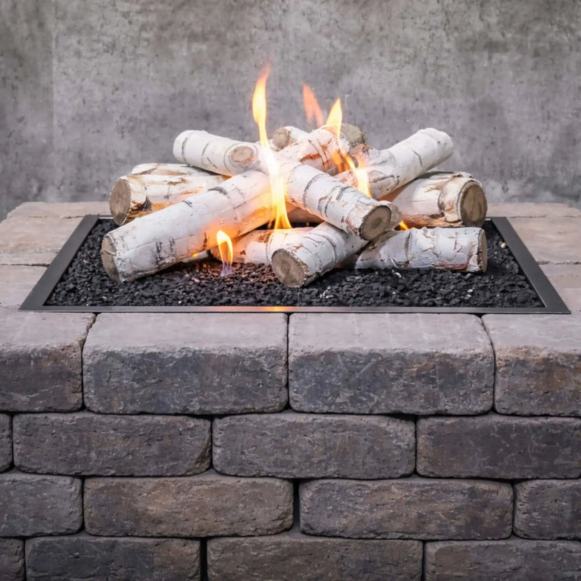 Close-up view of Sierra Birch ceramic gas fire pit logs arranged over black lava rock in a rectangular burner pan, with yellow flames rising between the white birch-style logs and a stacked stone fire pit wall beneath.