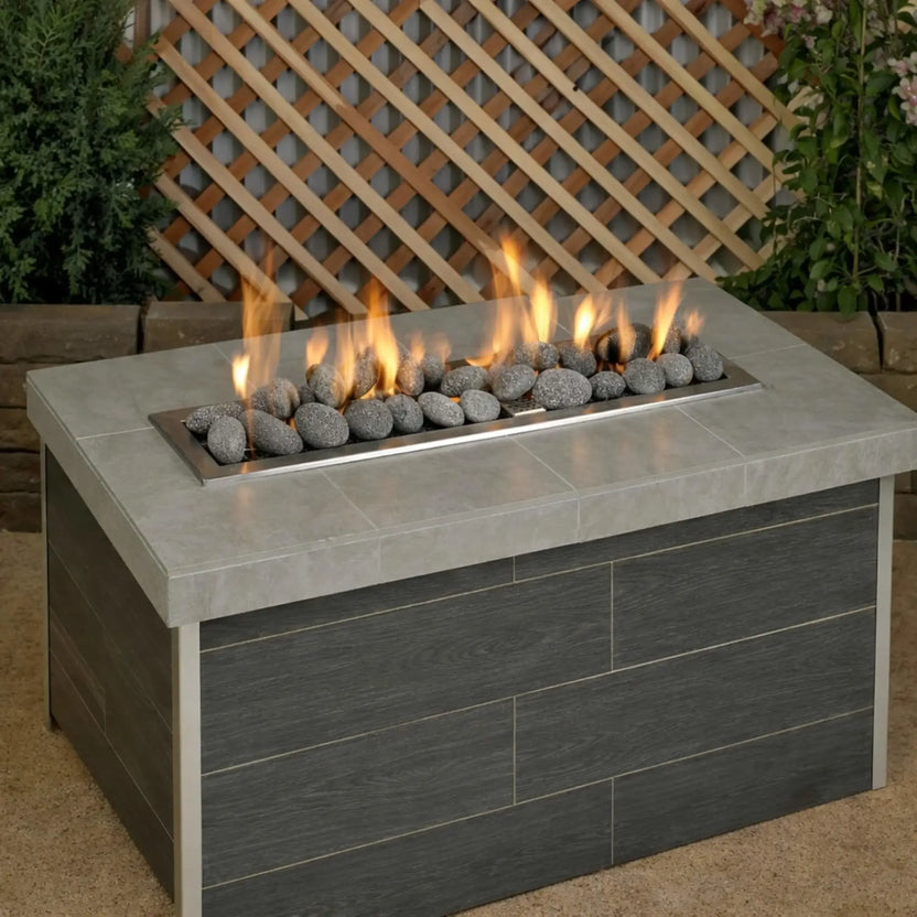 Modern rectangular outdoor gas fire table filled with Large gray lava stones arranged along a stainless steel linear burner, with steady yellow flames set against a backyard lattice fence and greenery.