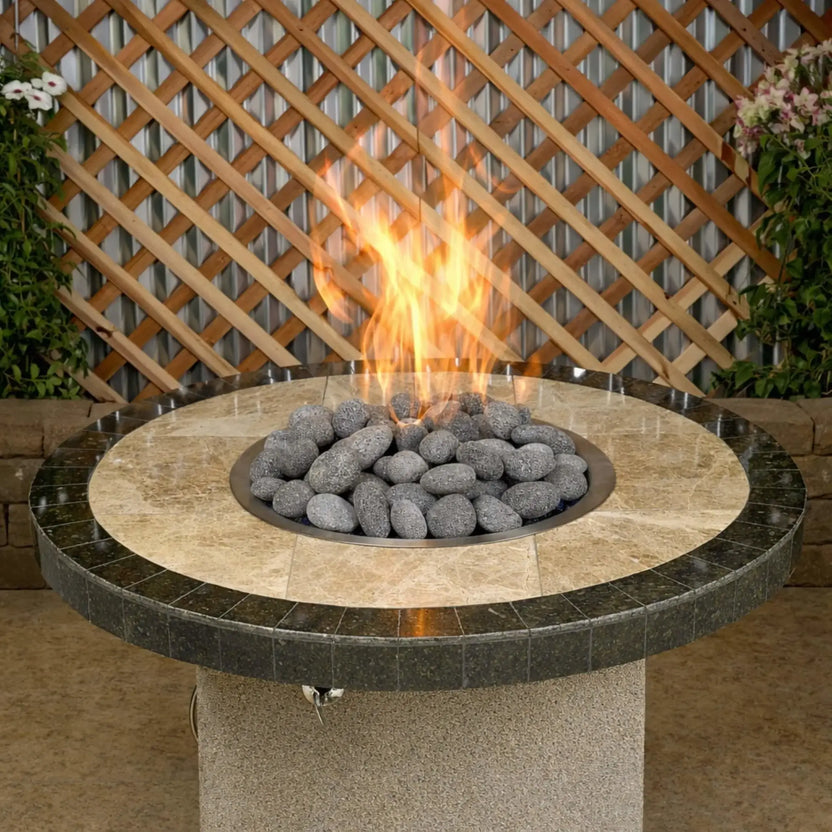 Round patio fire pit table featuring Large lava stones surrounding a central gas flame, displayed in an outdoor setting with a stone top, wood lattice backdrop, and landscaped plants.