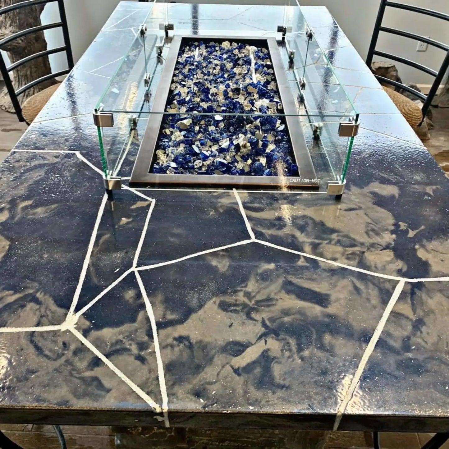Linear rectangular fire pit with a tempered glass wind guard installed on a stone countertop, featuring blue and clear fire glass inside a stainless steel burner pan in an indoor showroom setting.