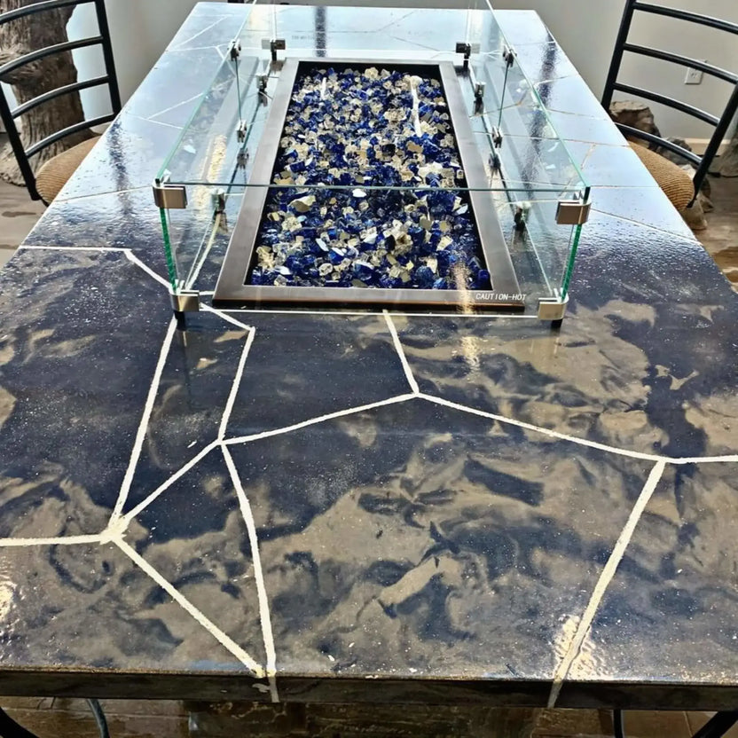 Linear rectangular fire pit with a tempered glass wind guard installed on a stone countertop, featuring blue and clear fire glass inside a stainless steel burner pan in an indoor showroom setting.