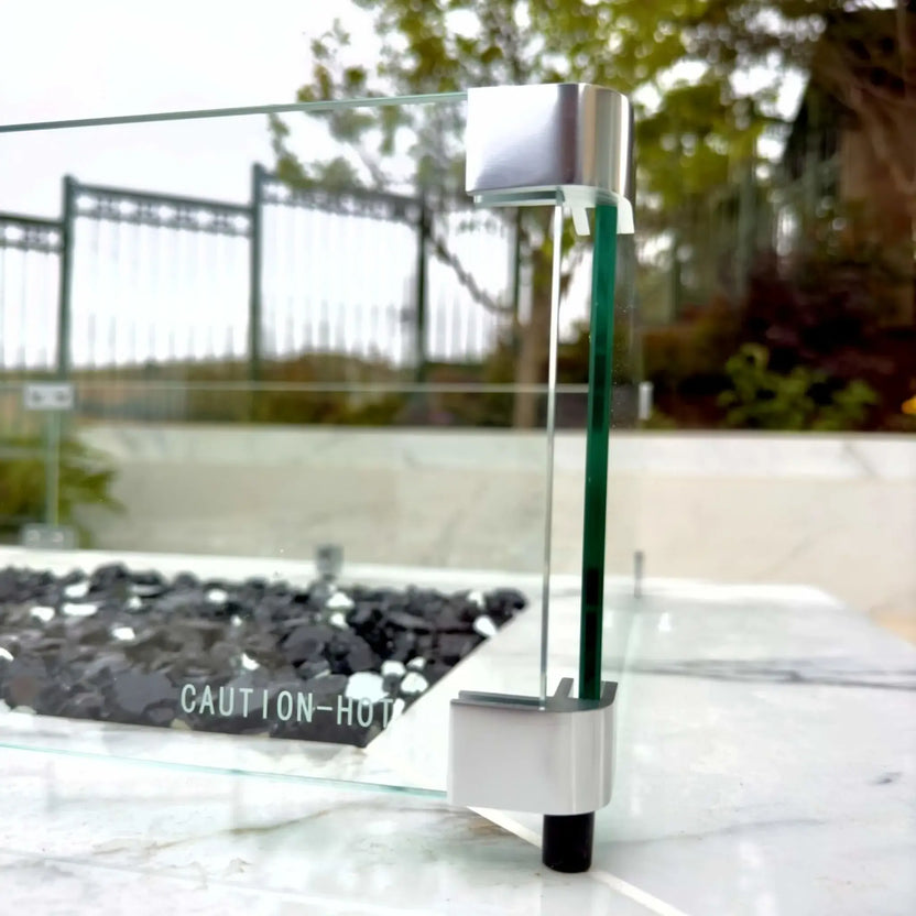 Detailed corner view of a tempered glass wind guard mounted on a linear fire pit, showing stainless steel brackets, clear glass panels, and black fire glass media inside a polished stone fire pit surround.
