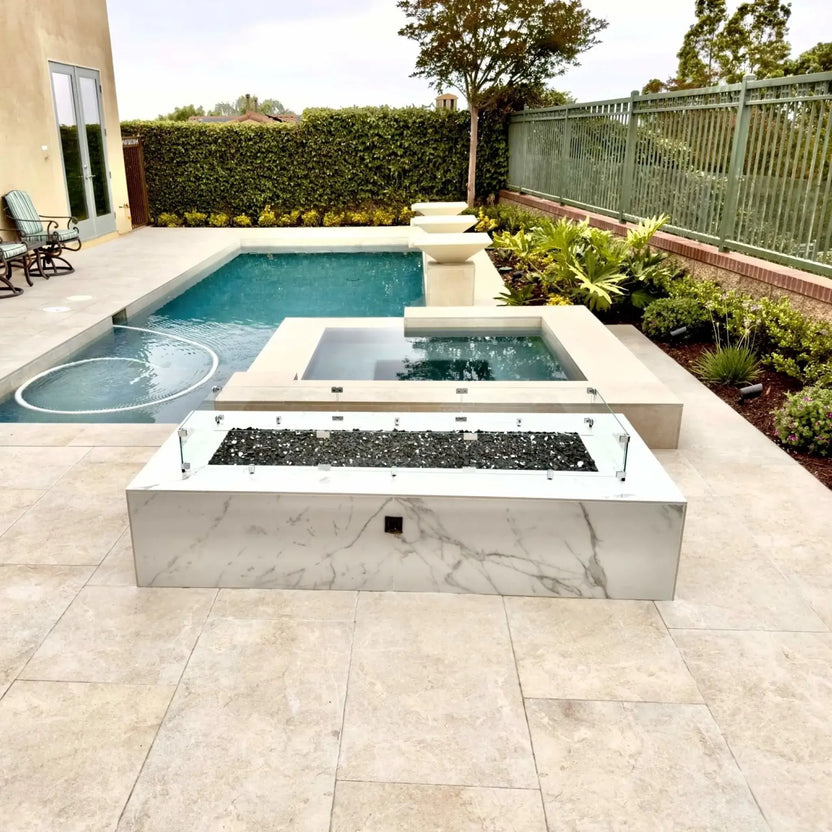 Contemporary backyard fire pit with a linear rectangular burner protected by a tempered glass wind guard, positioned near a swimming pool and spa, blending fire and water elements in a luxury outdoor living space.