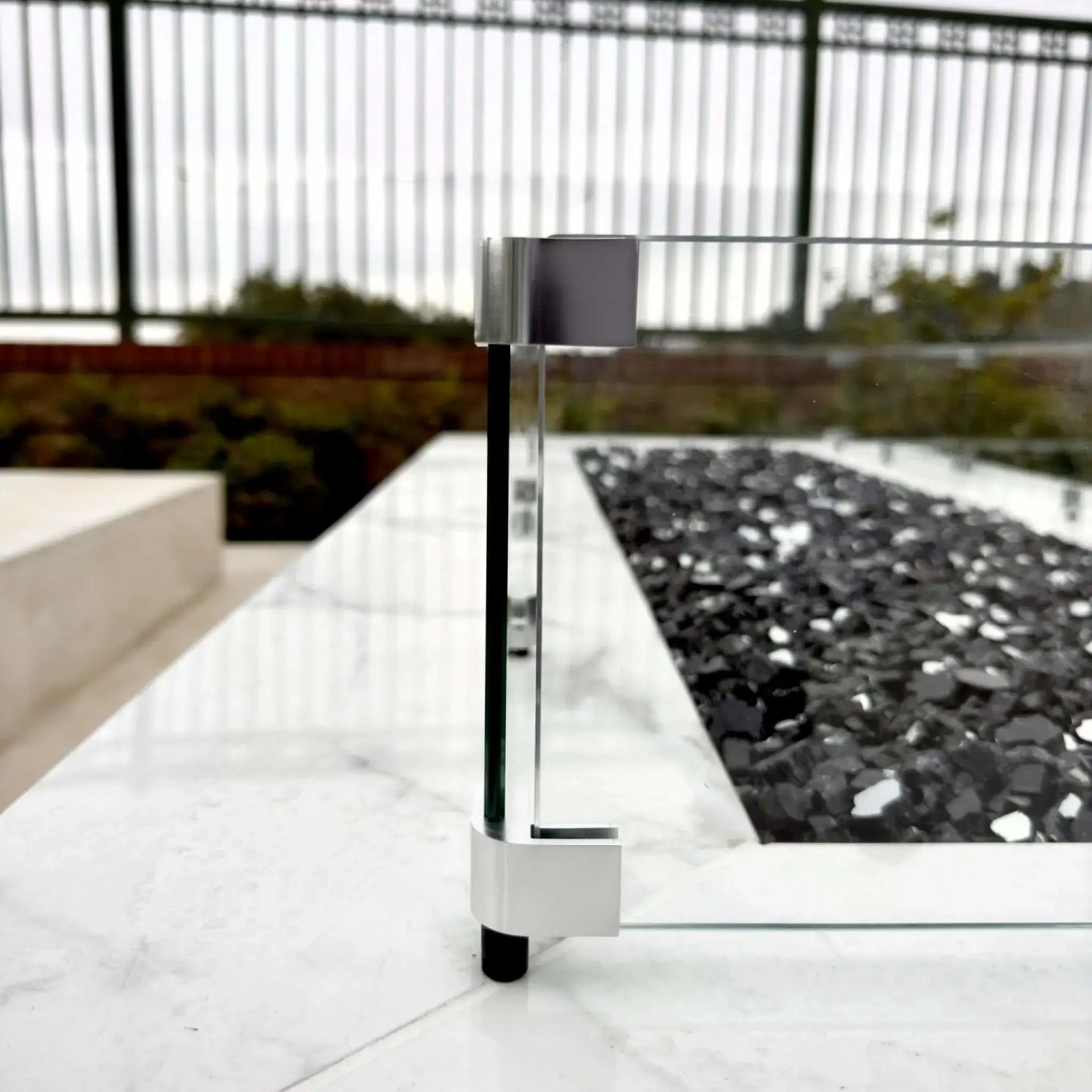 Side profile of a linear fire pit with a clear tempered glass wind guard, showcasing the low-profile glass panels, metal mounting hardware, and black fire glass set into a marble-style fire pit surface.