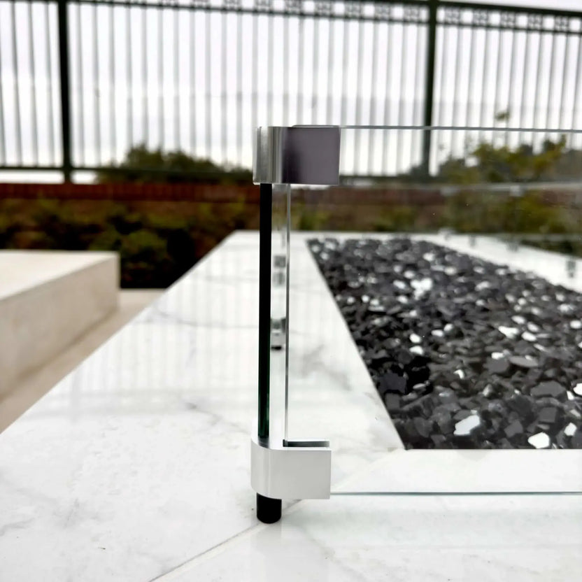 Side profile of a linear fire pit with a clear tempered glass wind guard, showcasing the low-profile glass panels, metal mounting hardware, and black fire glass set into a marble-style fire pit surface.