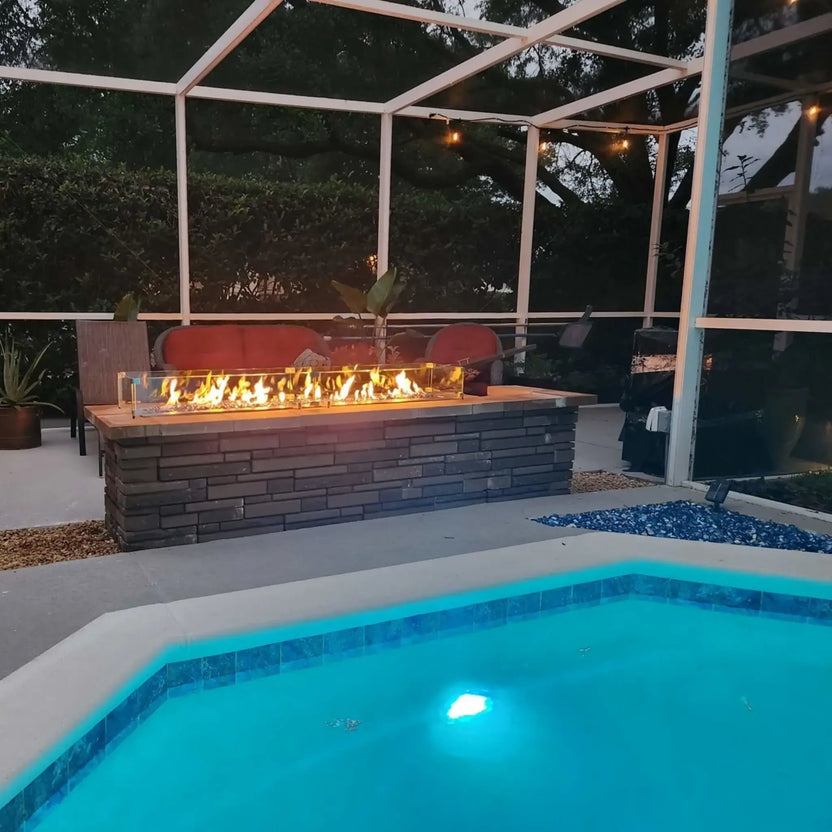 Evening poolside setting featuring a linear rectangular fire pit with active flames behind a tempered glass wind guard, installed in a stacked stone fire pit base beneath a screened patio enclosure.