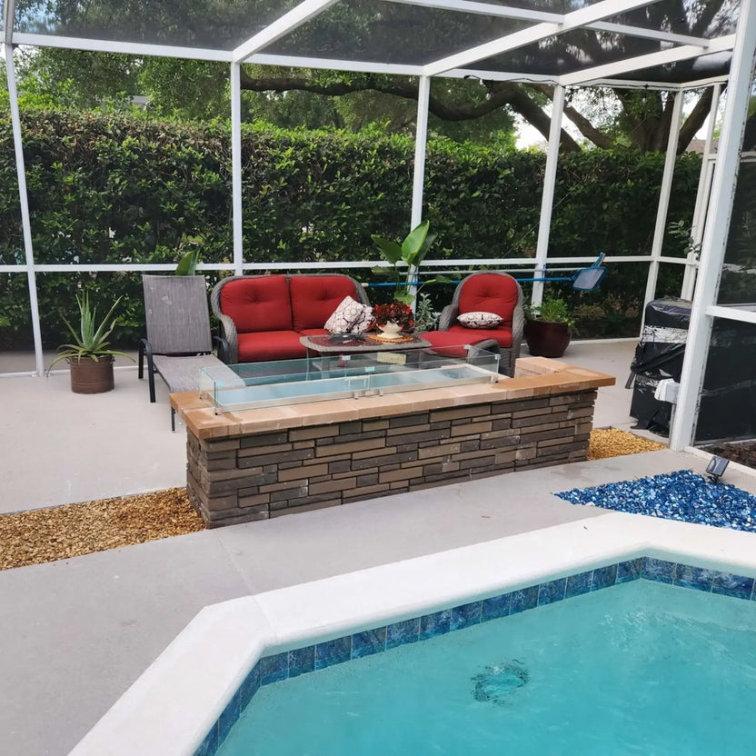 Backyard lounge area with cushioned seating and a modern linear fire pit topped with a tempered glass wind guard, positioned alongside a swimming pool to create a warm and inviting outdoor gathering space.