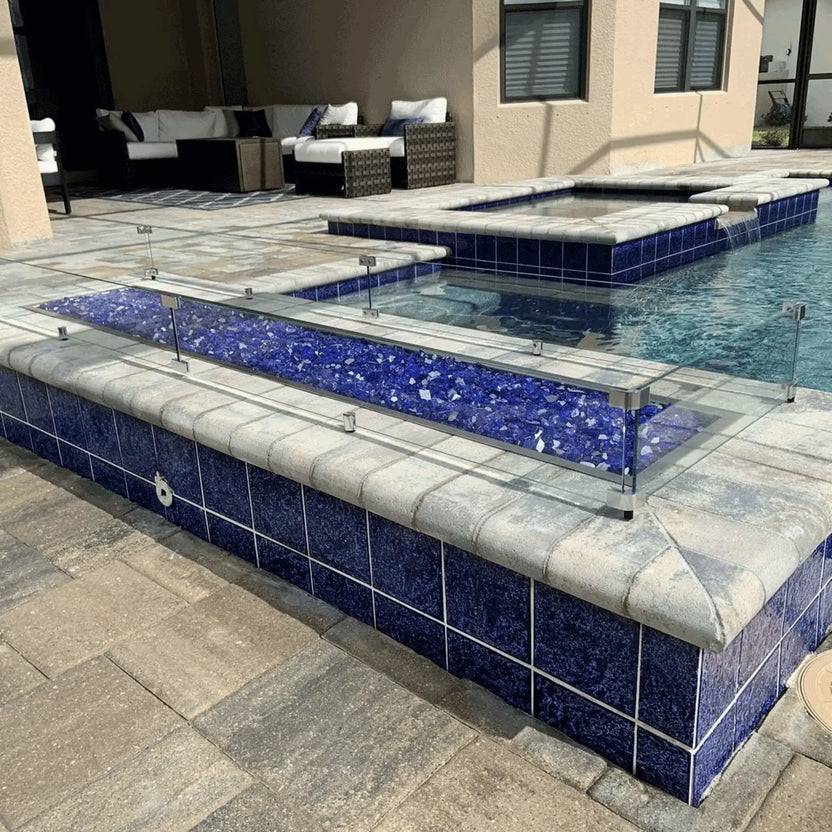 Linear rectangular fire pit installed along a raised pool edge, featuring a clear tempered glass wind guard and blue fire glass media set into a tiled outdoor pool and spa patio.