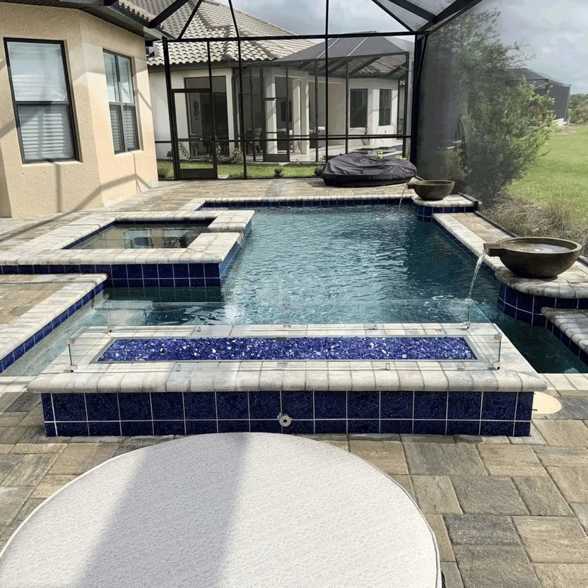 Modern backyard pool and spa with a linear fire pit accent, showcasing a tempered glass wind guard surrounding blue fire glass, integrated into a screened lanai outdoor living space.