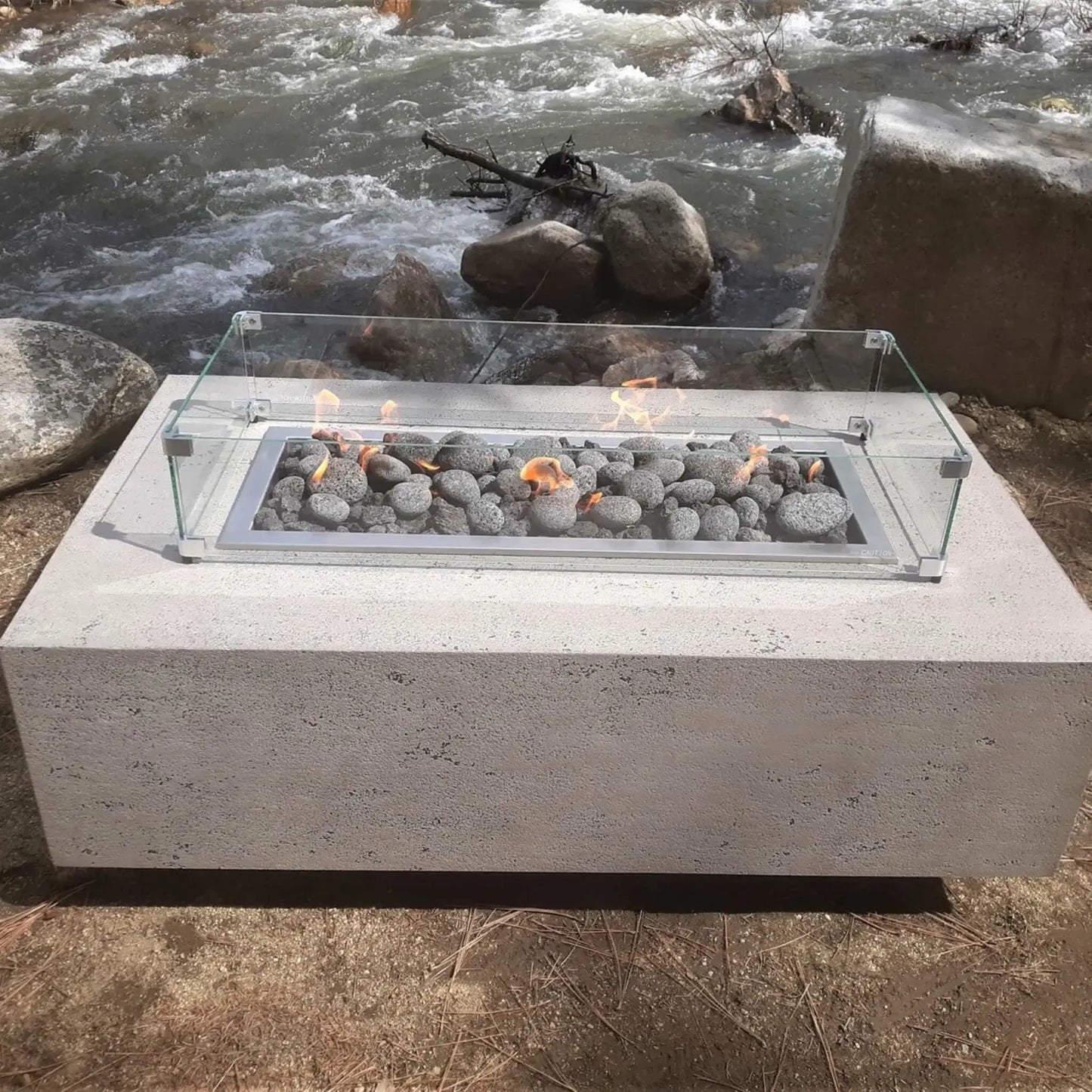 Concrete fire pit with a linear burner filled with smooth lava stones, protected by a clear glass wind guard, positioned beside a flowing river and natural rock landscape.