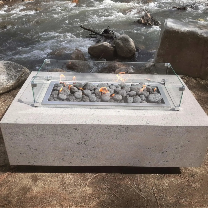 Concrete fire pit with a linear burner filled with smooth lava stones, protected by a clear glass wind guard, positioned beside a flowing river and natural rock landscape.