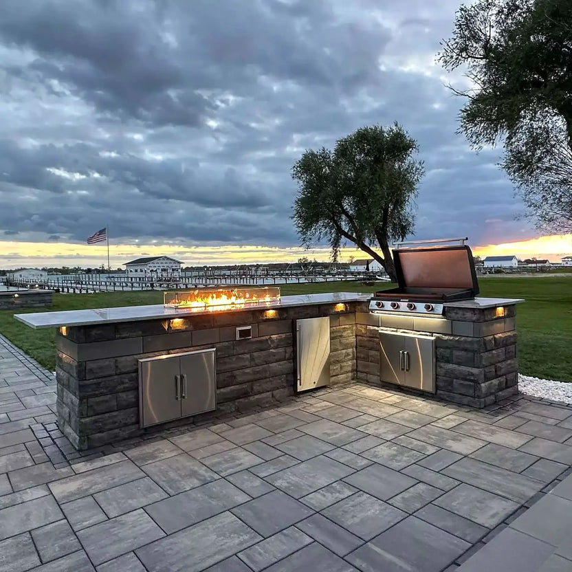 Outdoor kitchen island with a built-in linear fire feature, topped with a glass wind guard and decorative fire glass, set against a waterfront backdrop at sunset.