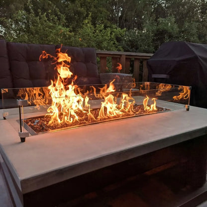 Close-up view of a linear fire pit actively burning with tall flames, framed by a tempered glass wind guard and installed on a contemporary concrete fire table in a backyard lounge area.