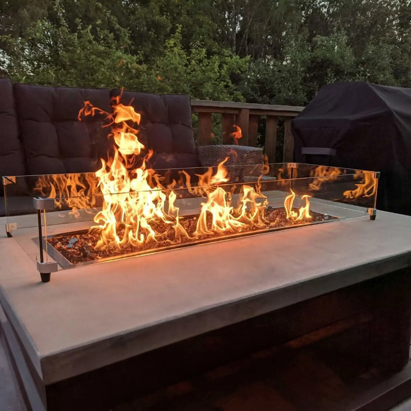 Close-up view of a linear fire pit actively burning with tall flames, framed by a tempered glass wind guard and installed on a contemporary concrete fire table in a backyard lounge area.