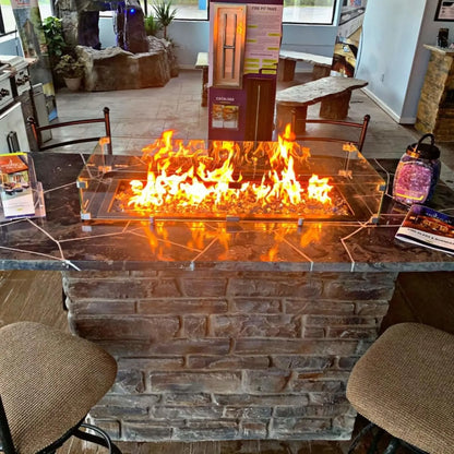 Modern linear fire pit burning with bright orange flames, protected by a clear tempered glass wind guard, set into a stone island with bar seating inside a fire feature showroom.