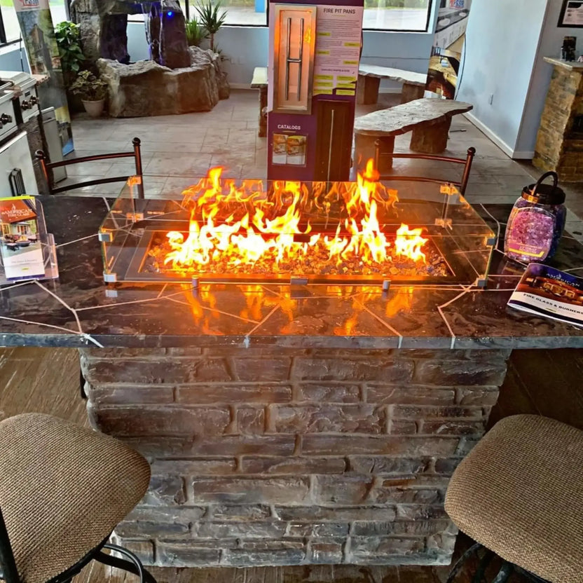 Modern linear fire pit burning with bright orange flames, protected by a clear tempered glass wind guard, set into a stone island with bar seating inside a fire feature showroom.