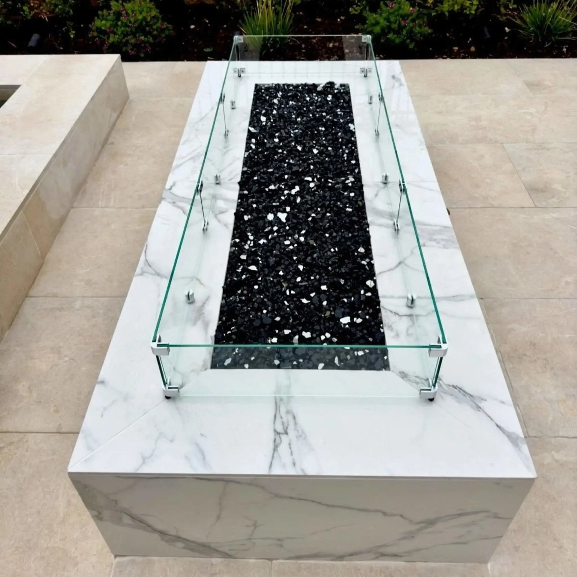Top-down view of a rectangular fire pit featuring black fire glass media, surrounded by a clear tempered glass wind guard mounted on a polished stone fire table.