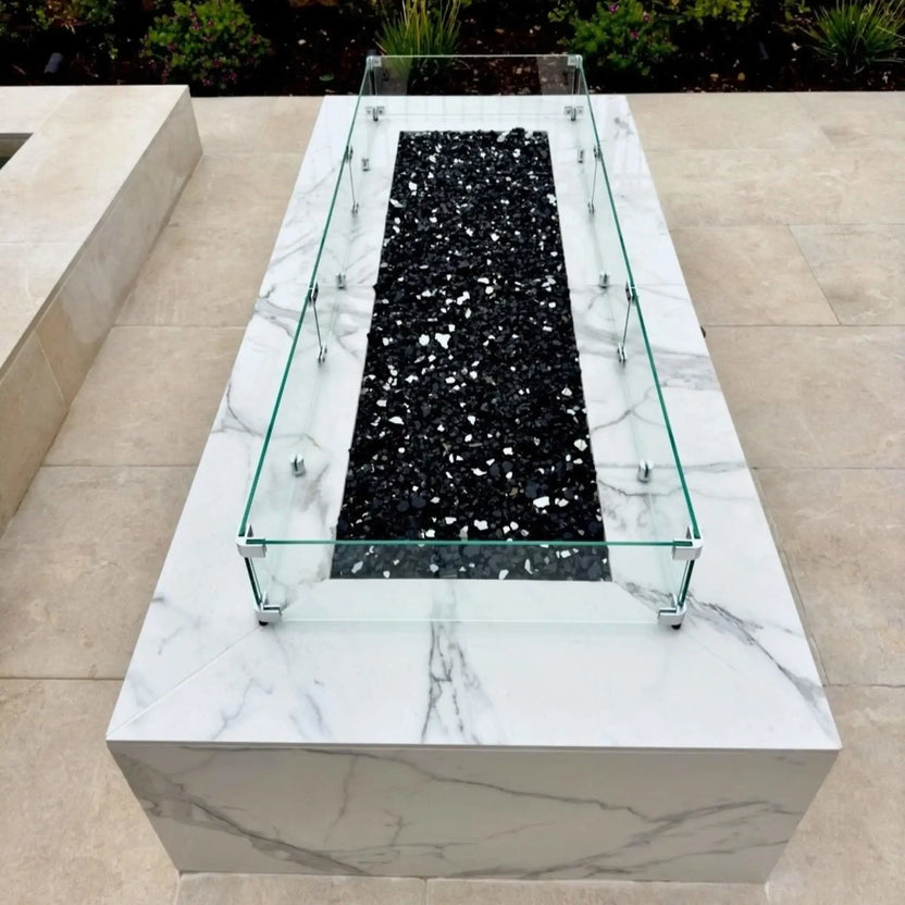 Top-down view of a rectangular fire pit featuring black fire glass media, surrounded by a clear tempered glass wind guard mounted on a polished stone fire table.