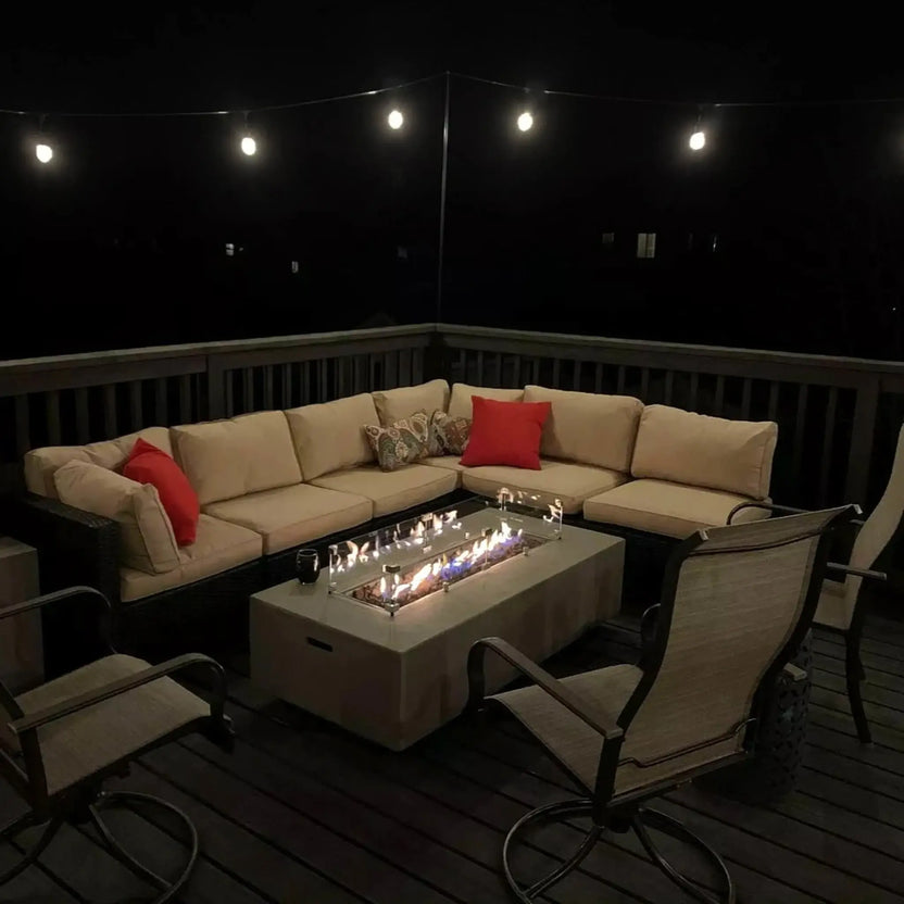 Evening patio setting with a linear fire pit centerpiece, protected by a glass wind guard and glowing fire glass, surrounded by cushioned outdoor seating and string lights on a wooden deck.