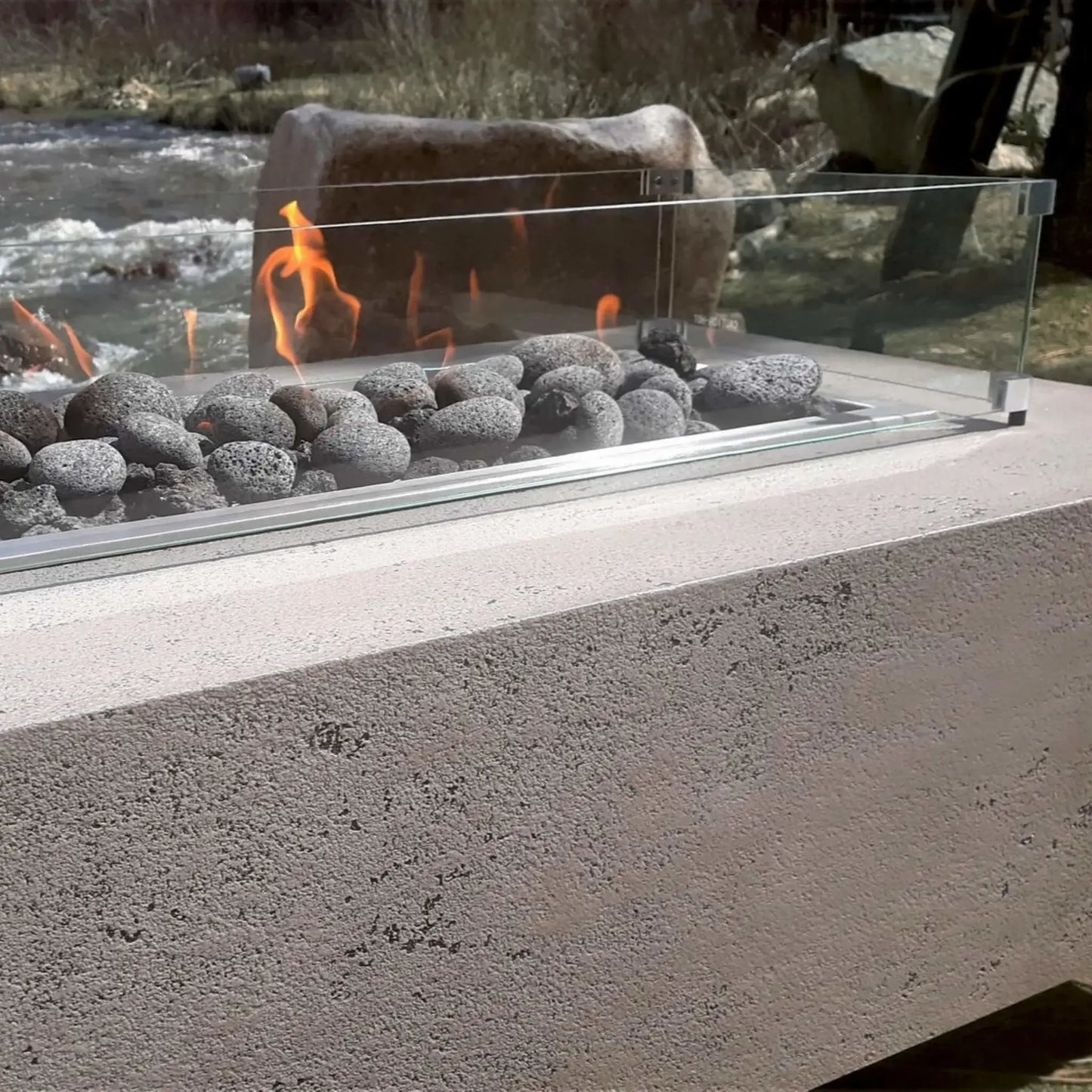 Linear rectangular gas fire pit installed beside a flowing river, featuring smooth gray lava stones and a clear tempered glass wind guard that shields the flame while maintaining unobstructed views of the surrounding natural landscape.