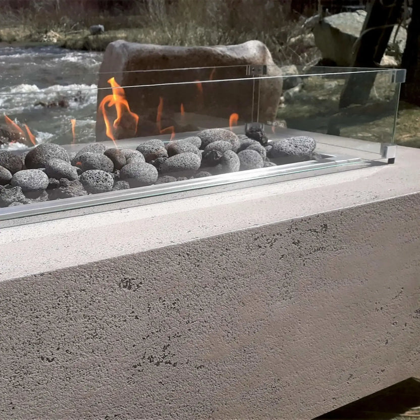 Linear rectangular gas fire pit installed beside a flowing river, featuring smooth gray lava stones and a clear tempered glass wind guard that shields the flame while maintaining unobstructed views of the surrounding natural landscape.