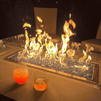 Nighttime outdoor fire pit scene with a linear gas burner filled with blue fire glass, protected by a clear glass wind guard, set on a modern patio table with two drinks placed nearby for a relaxed evening ambiance.