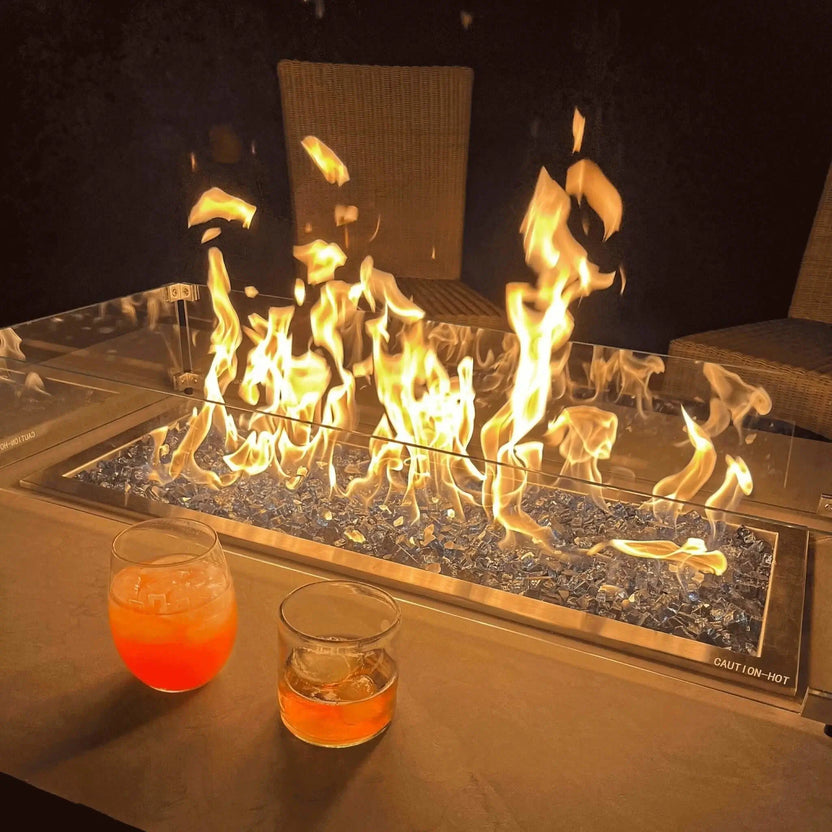 Nighttime outdoor fire pit scene with a linear gas burner filled with blue fire glass, protected by a clear glass wind guard, set on a modern patio table with two drinks placed nearby for a relaxed evening ambiance.