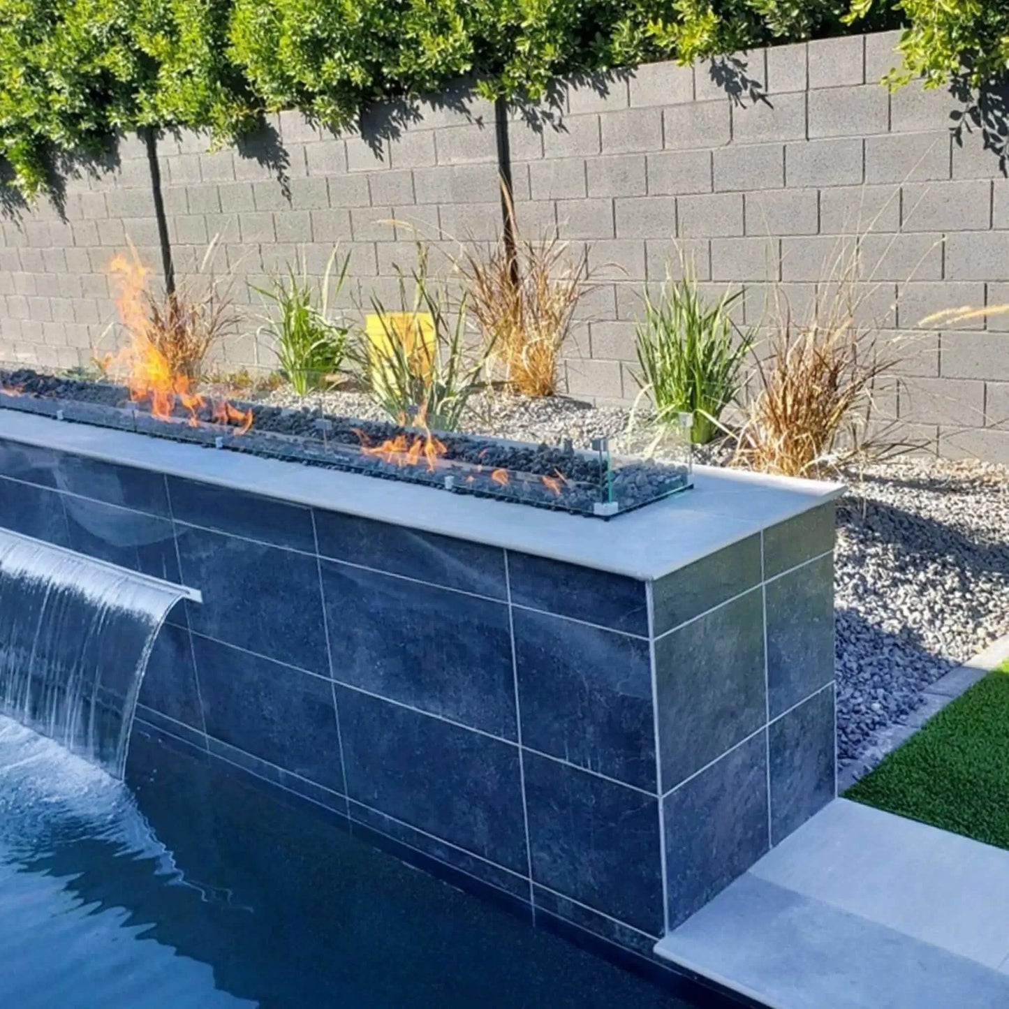 Contemporary poolside fire feature with a long linear gas fire pit mounted on a raised tiled wall, showcasing dancing flames over fire media and enclosed by a tempered glass wind guard for wind protection and safety.