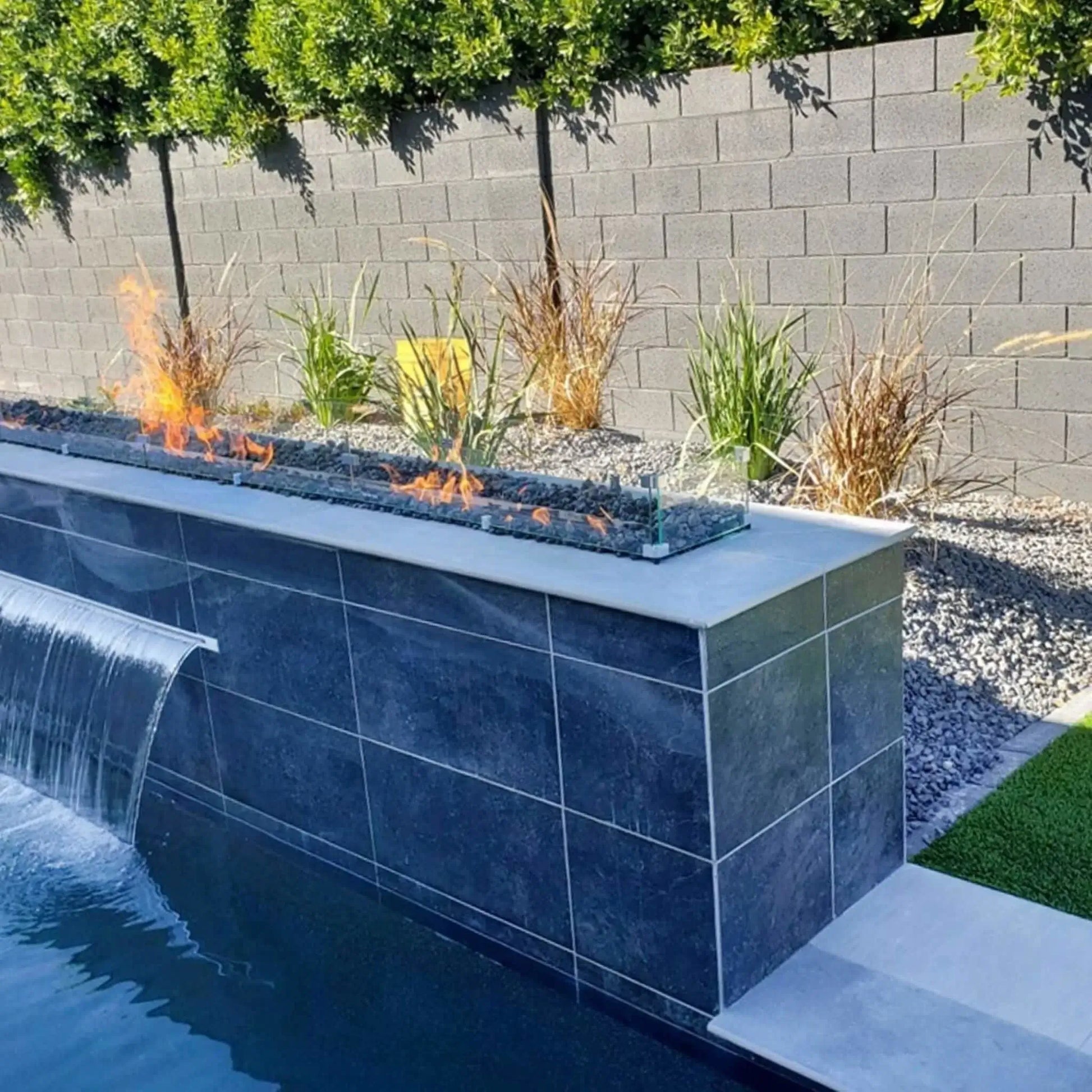 Contemporary poolside fire feature with a long linear gas fire pit mounted on a raised tiled wall, showcasing dancing flames over fire media and enclosed by a tempered glass wind guard for wind protection and safety.