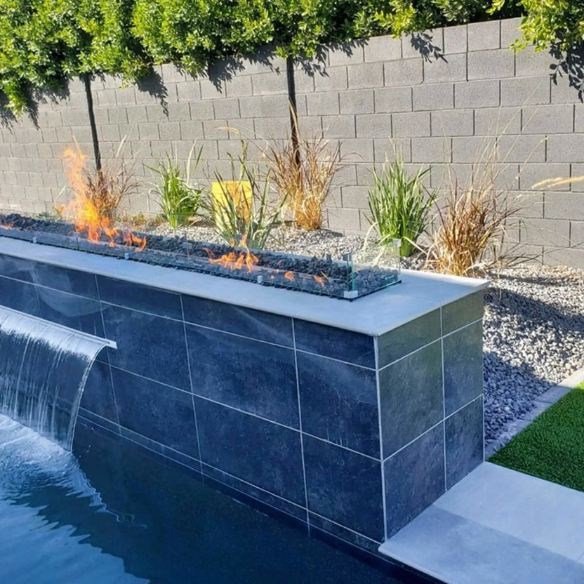 Contemporary poolside fire feature with a long linear gas fire pit mounted on a raised tiled wall, showcasing dancing flames over fire media and enclosed by a tempered glass wind guard for wind protection and safety.
