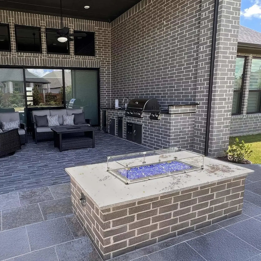 Covered outdoor patio with a brick fire pit island topped by a linear gas burner and blue fire glass, surrounded by a low-profile tempered glass wind guard and positioned near an outdoor kitchen and seating area.