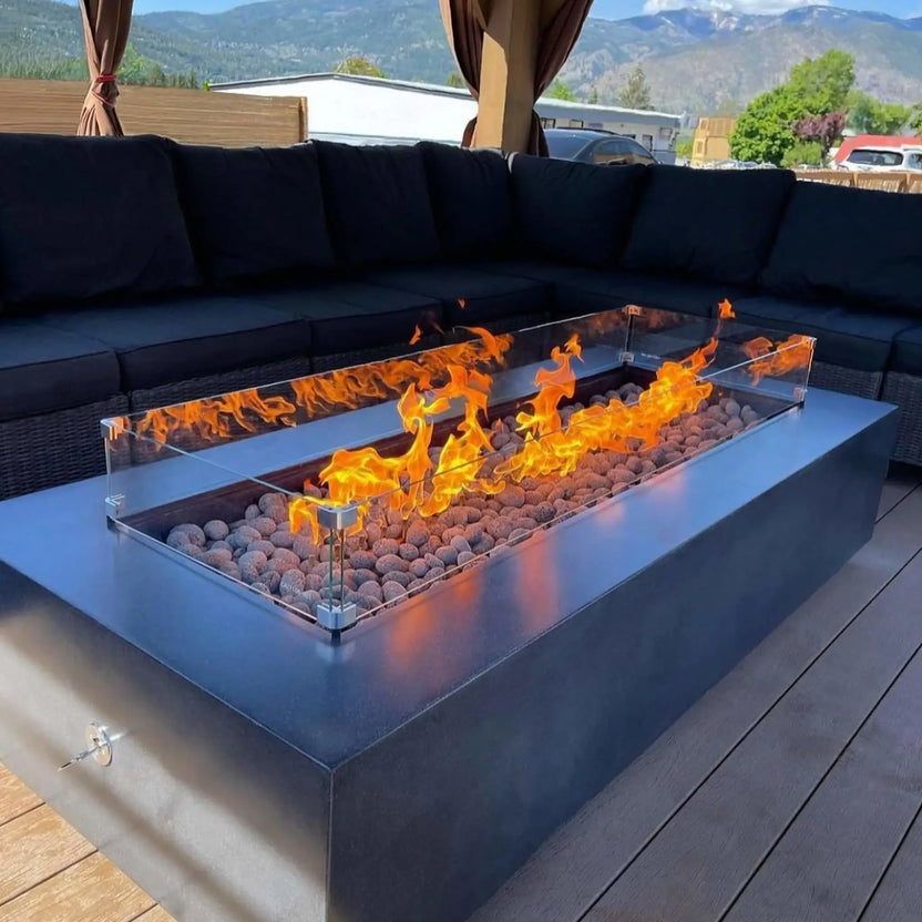 Modern outdoor fire pit with a matte black rectangular enclosure, filled with round lava stones and enclosed by a clear glass wind guard, placed on a deck with sectional seating and scenic mountain views in the background.