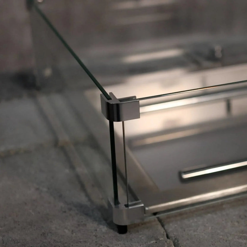 Close-up detail of a tempered glass wind guard corner bracket on a linear fire pit, showing brushed metal hardware securely holding the glass panels above a paved outdoor surface.