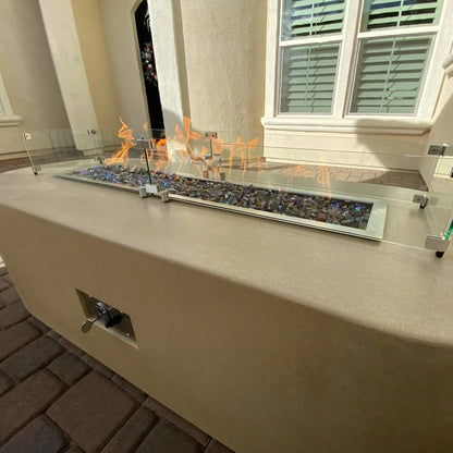 Linear outdoor gas fire pit built into a stucco fire table, featuring multicolor fire glass, visible flames, and a surrounding clear glass wind guard, installed in a residential courtyard with paver flooring.