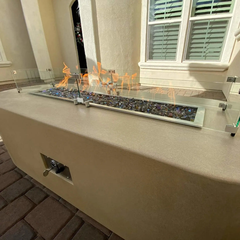Linear outdoor gas fire pit built into a stucco fire table, featuring multicolor fire glass, visible flames, and a surrounding clear glass wind guard, installed in a residential courtyard with paver flooring.