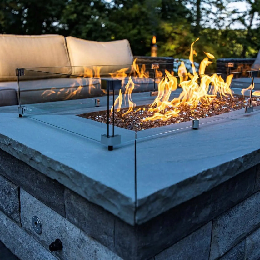 Linear rectangular outdoor fire pit with a clear tempered glass wind guard, featuring bright flames dancing over fire glass, set into a stone fire pit table beside cushioned patio seating in a landscaped backyard.
