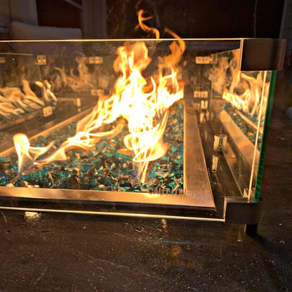 Close-up view of a linear fire pit with active flames dancing over blue fire glass, enclosed by a clear tempered glass wind guard secured with metal brackets on a stone surface.