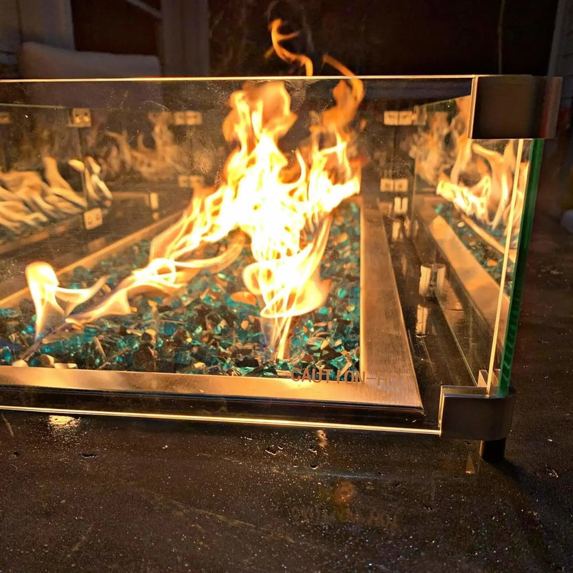 Close-up view of a linear fire pit with active flames dancing over blue fire glass, enclosed by a clear tempered glass wind guard secured with metal brackets on a stone surface.