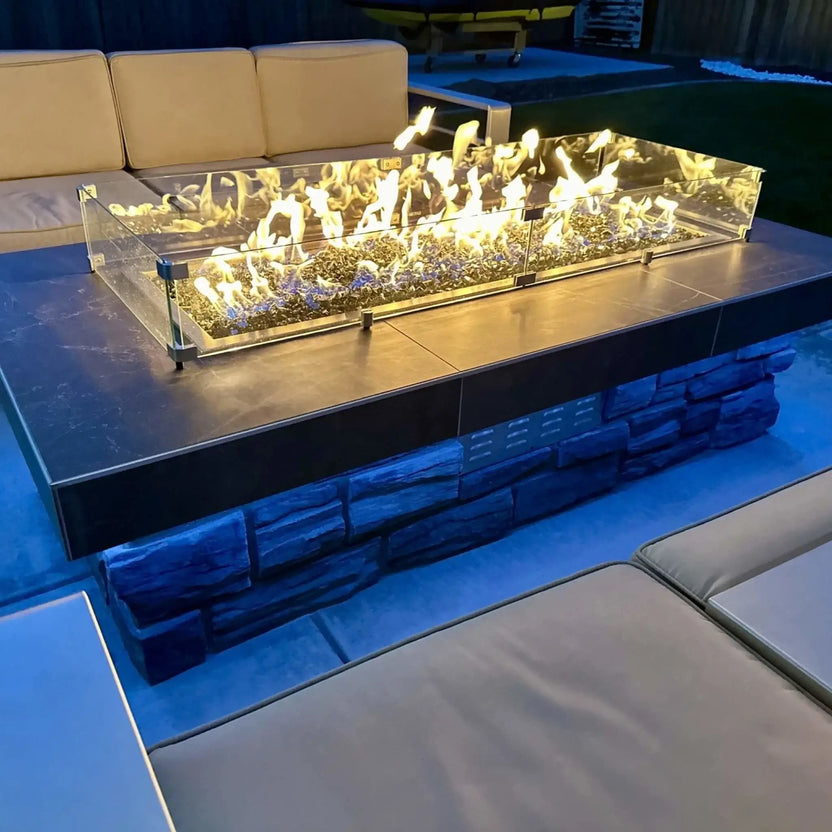 Modern linear fire pit with tempered glass wind guard installed on a raised stone base, showcasing vibrant flames over decorative fire glass, surrounded by contemporary outdoor lounge furniture at dusk.
