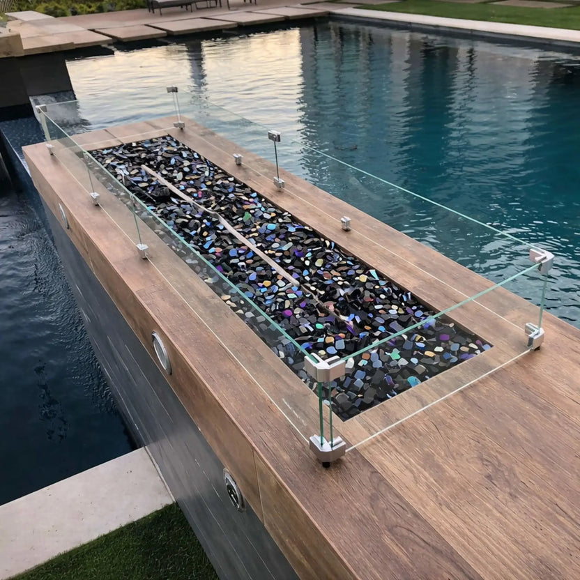 Rectangular fire pit integrated into a poolside water feature, topped with a clear glass wind guard and filled with reflective fire glass, highlighting a sleek outdoor design next to a swimming pool.