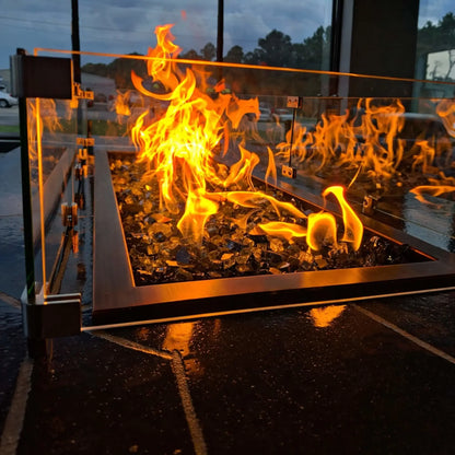 Close-up view of a linear fire pit with tempered glass wind guard, showing tall orange flames rising above fire glass, framed by stainless steel trim in a modern outdoor setting.