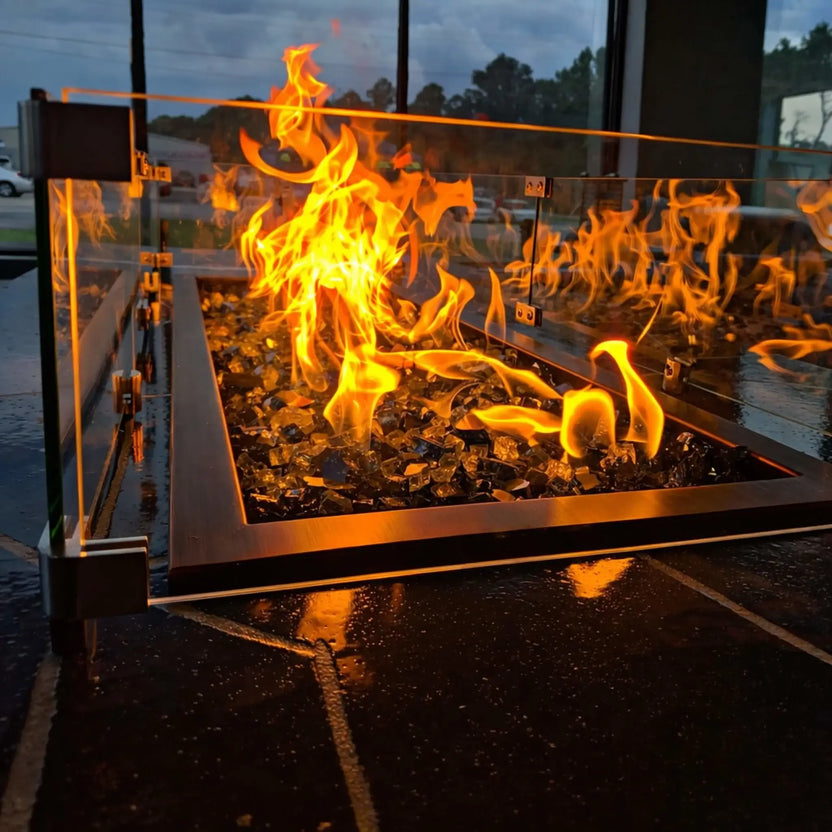 Close-up view of a linear fire pit with tempered glass wind guard, showing tall orange flames rising above fire glass, framed by stainless steel trim in a modern outdoor setting.