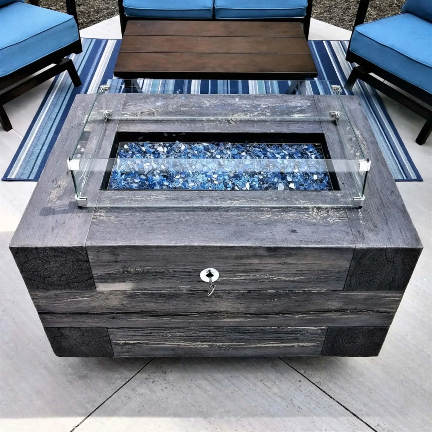 Rectangular fire pit table with clear glass wind guard and multicolored fire glass, set between cushioned patio chairs on a stone patio, creating a cozy outdoor gathering space.