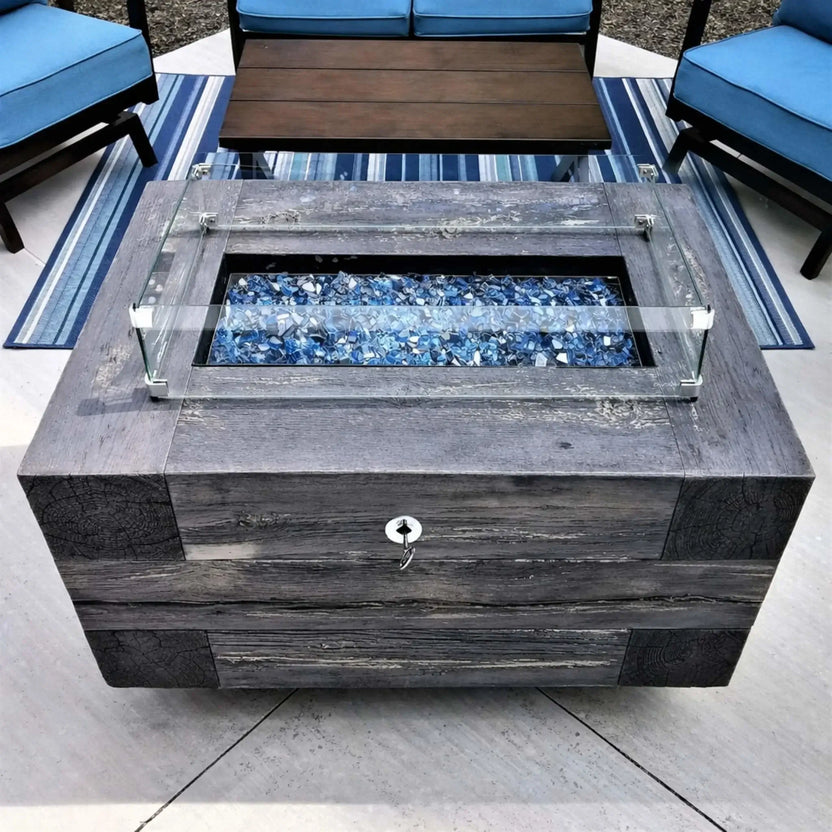 Rectangular fire pit table with clear glass wind guard and multicolored fire glass, set between cushioned patio chairs on a stone patio, creating a cozy outdoor gathering space.