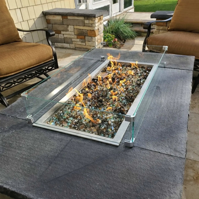 Outdoor linear fire pit with tempered glass wind guard installed in a stone fire pit table, featuring steady flames over fire glass, surrounded by classic patio furniture and landscaped stonework.