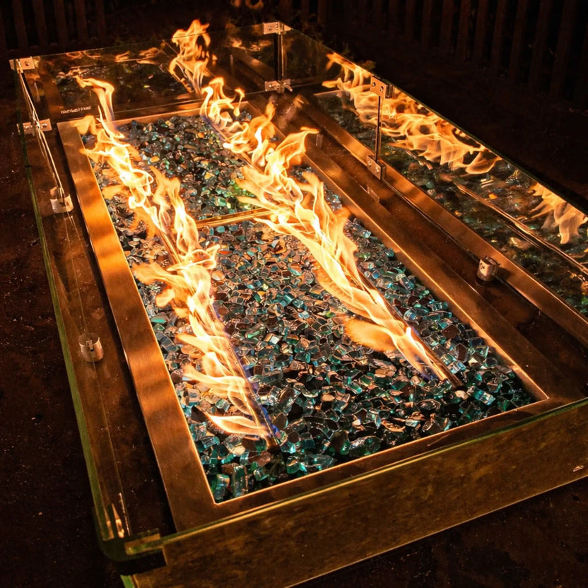 Dramatic nighttime view of a linear fire pit with tempered glass wind guard, showcasing bright flames reflecting off fire glass and glass panels, emphasizing a modern outdoor fire feature design.