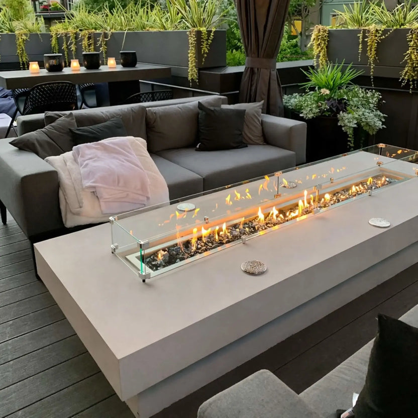 Modern outdoor patio seating area featuring a long linear fire pit table with a tempered glass wind guard, glowing flames over fire glass, and gray cushioned sofas surrounded by raised planters and soft ambient lighting.