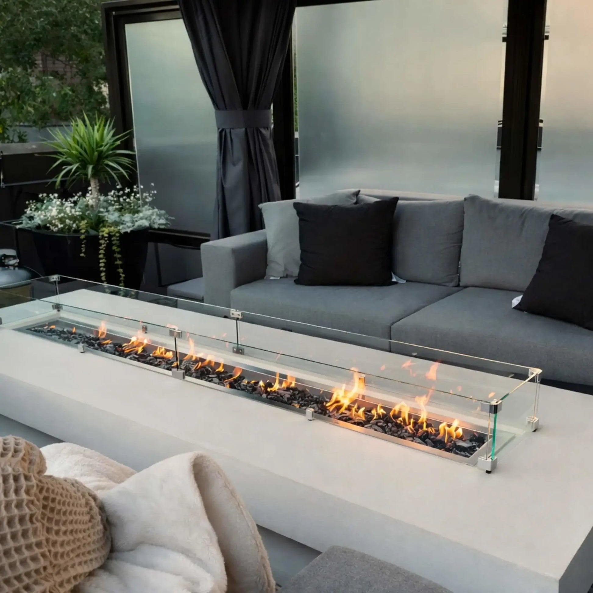 Contemporary backyard lounge with a sleek rectangular fire pit protected by a clear glass wind guard, bright flames dancing over dark fire glass, and deep gray outdoor couches framed by privacy panels and greenery.