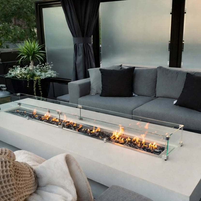 Contemporary backyard lounge with a sleek rectangular fire pit protected by a clear glass wind guard, bright flames dancing over dark fire glass, and deep gray outdoor couches framed by privacy panels and greenery.