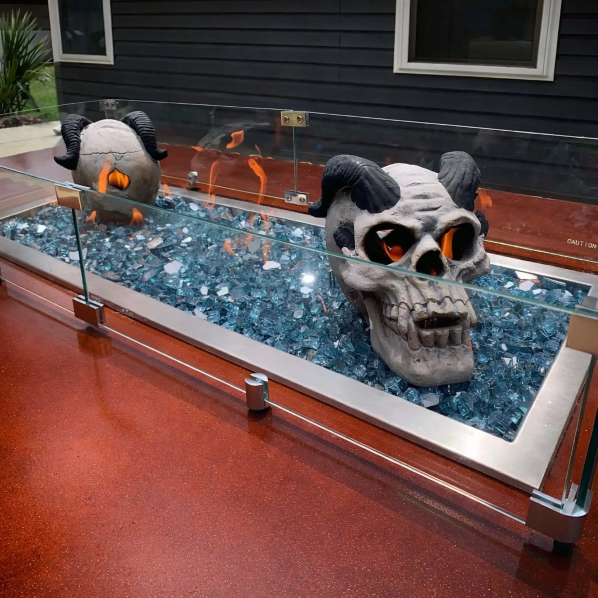 Linear fire pit installation filled with blue fire glass and decorative skull accents, enclosed by a tempered glass wind guard, creating a bold custom fire feature set into a red concrete patio surface.